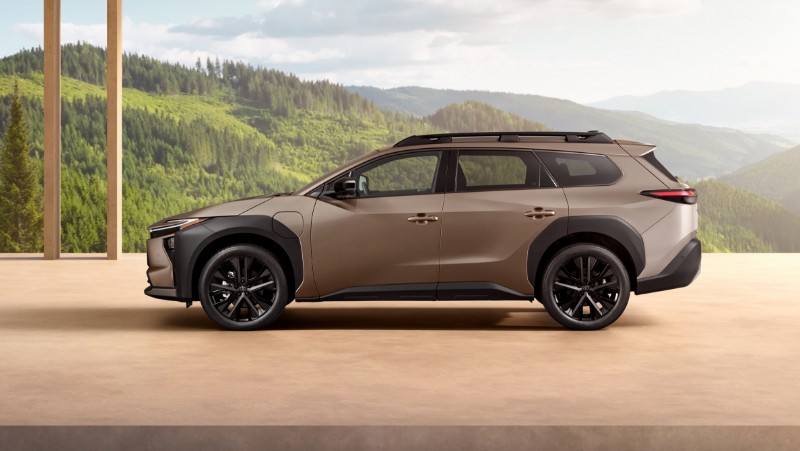 Profile of a bronze Toyota bZ4X Touring SUV parked on a wooden floor. A mountain and forest landscape visible in the background.
